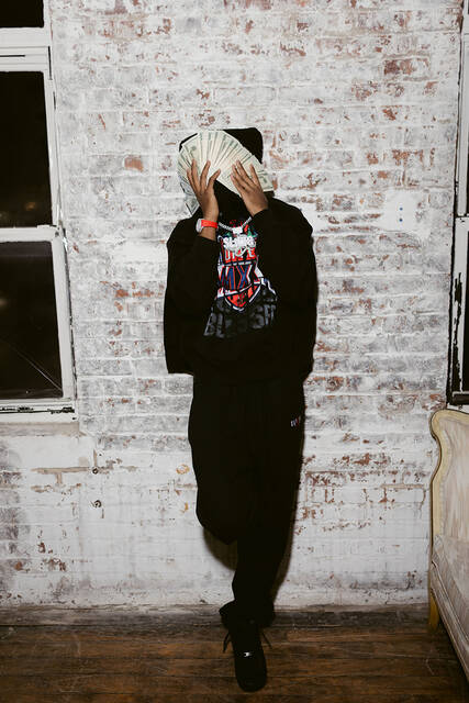 RodSlime holding a stack of cash over his face against a whitewashed brick wall.